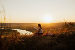 A young woman reading a book outdoors during a sunset.