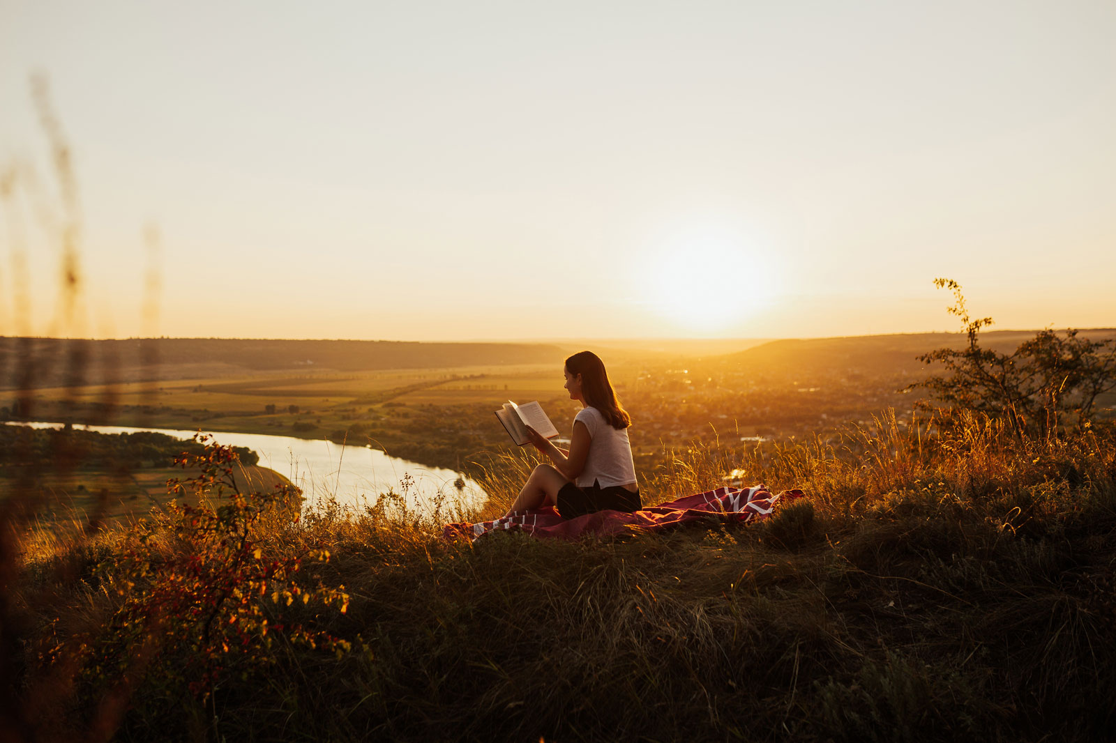 A young woman reading a book outdoors during a sunset.