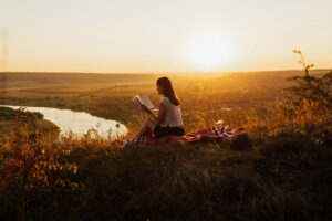 A photo of a young woman reading a book outdoors during a sunset.