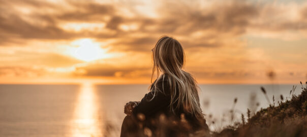 A woman sitting by the ocean at sunset looking out towards the horizon.